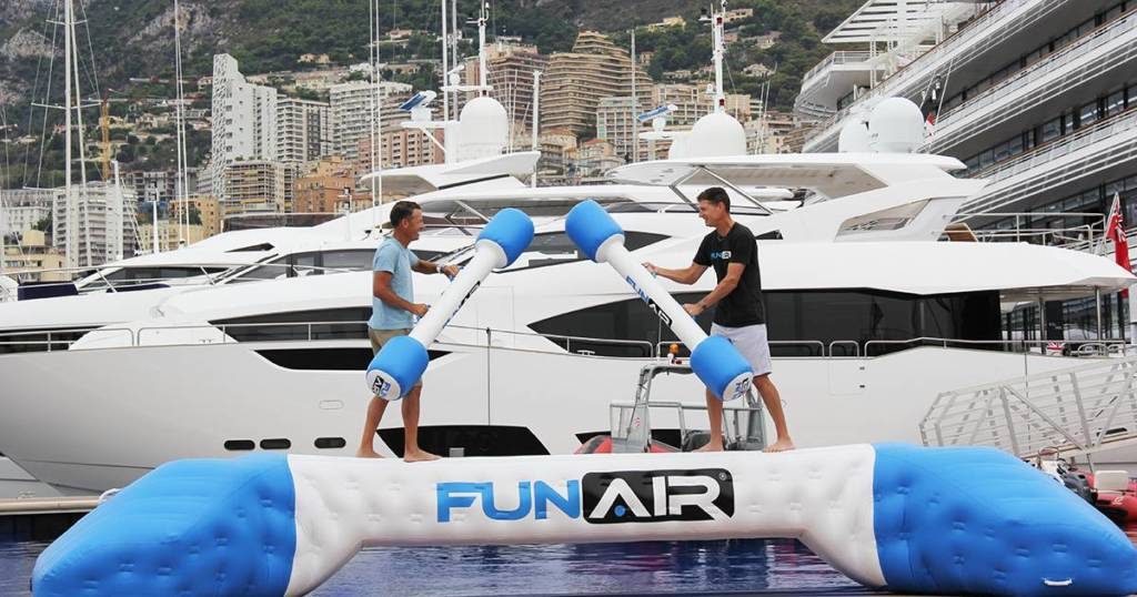 Two men stood on a FunAIr Water Joust Balance BEan about to battle with inflatable poles with a superyacht behind them